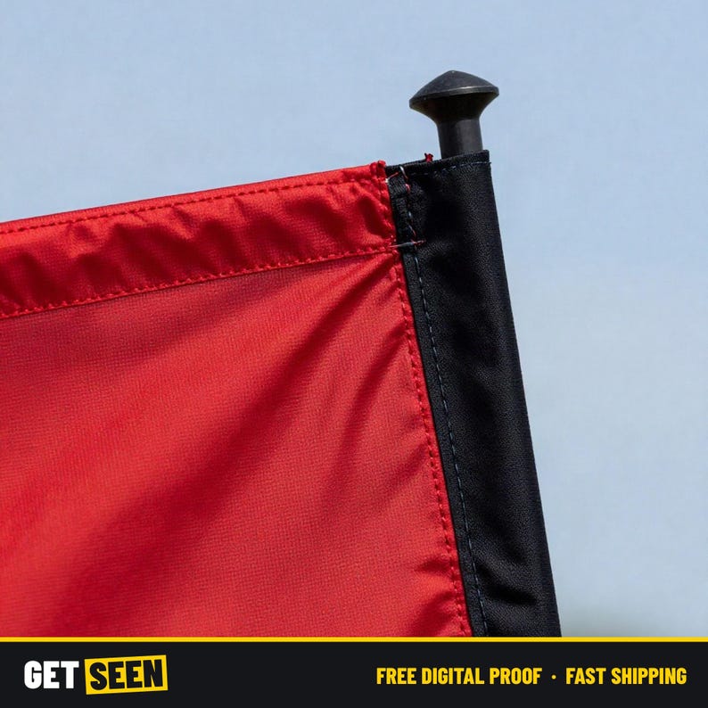 May include: A red flag with a black vertical sleeve attached to a black pole featuring a rounded top cap. The fabric has a visible stitched seam where the red material meets the black sleeve. The background is a solid light blue color.