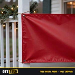 May include: A rectangular red banner made of smooth material featuring silver metal grommets along the edges. The banner is secured to a white vertical post with twine, positioned near green pine branches with small warm white lights.