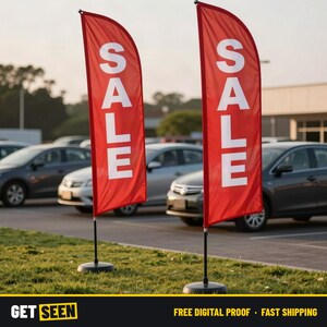 May include: Two tall, red feather-style advertising flags standing on black circular bases. Each flag features the word SALE printed in large, bold, white capital letters oriented vertically. The flags are positioned upright on a grassy area.
