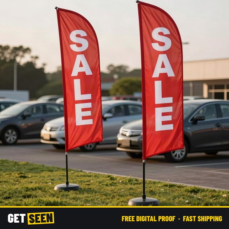 May include: Two tall, red feather-style advertising flags standing on black circular bases. Each flag features the word SALE printed in large, bold, white capital letters oriented vertically. The flags are positioned upright on a grassy area.