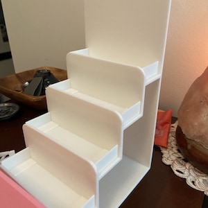 May include: A white acrylic display stand featuring four tiered shelves arranged in a staircase pattern. The structure includes a tall vertical back panel and four horizontal platforms with raised edges, designed for organising and showcasing items.