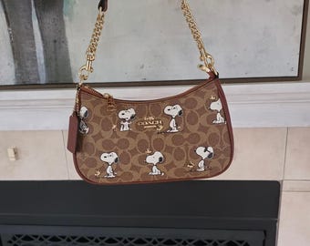 Puede incluir: Un bolso de hombro Coach marrón con un patrón de logotipo repetitivo y un estampado de Snoopy y Woodstock. El bolso tiene una correa de cadena dorada, ribetes de cuero marrón, cierre de cremallera superior y una etiqueta de cuero colgante.