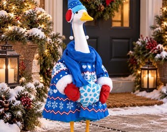 Op de afbeelding: Een wit ganzenbeeldje met een blauw-witte trui met sneeuwvlokpatronen, een blauwe sjaal, rode wanten en een blauwe muts met rode oorkappen. De gans houdt een blauw-wit kussen in de vorm van een sneeuwvlok vast in de vleugels.