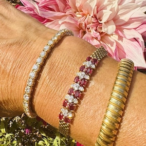 Puede incluir: Tres pulseras usadas juntas: una pulsera de tenis dorada con piedras redondas transparentes, una pulsera dorada con una doble fila de piedras ovaladas rojas acentuadas con pequeñas piedras transparentes y una pulsera rígida dorada acanalada.