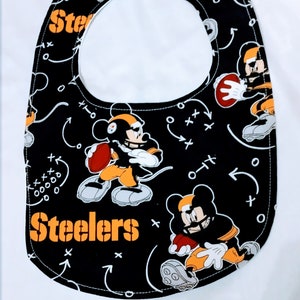 May include: Black baby bib with a Steelers football theme. The bib features cartoon characters in Steelers uniforms, footballs, and play diagrams. The word "Steelers" is printed in orange.