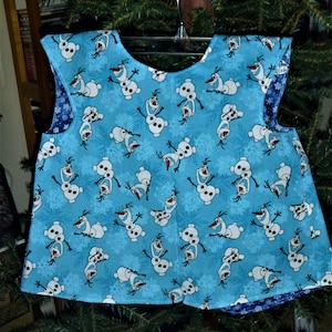 May include: A reversible sleeveless top featuring a light blue fabric with a pattern of Olaf the snowman from Frozen and snowflakes. The reverse side is a dark blue fabric with white snowflakes. The top is hanging on a hanger.