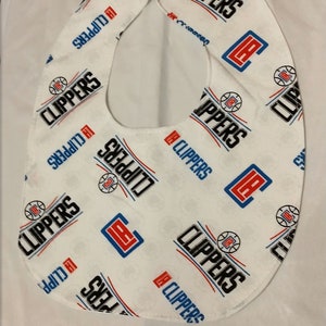 May include: White baby bib with a repeating pattern of the Los Angeles Clippers logo and team name in black, red, and blue. The bib has a curved shape with a neck opening and is made of soft fabric.