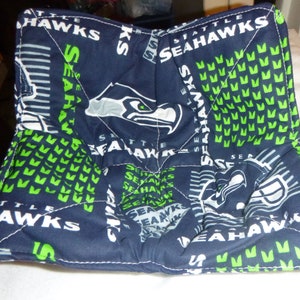 May include: A handmade fabric bowl cozy featuring a navy blue and green Seattle Seahawks print. The bowl has a square shape with white stitching and is designed to hold a bowl of hot food. The Seahawks logo and team name are visible.