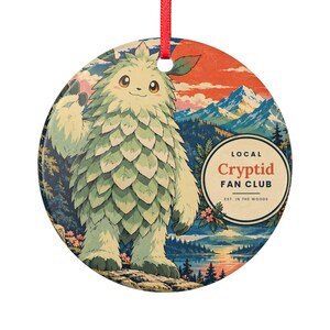 Op de afbeelding: Een rond keramisch ornament met een harig, lichtgroen wezen met bladvormige vacht in een berglandschap. De tekst luidt LOCAL Cryptid FAN CLUB EST. IN THE WOODS. Een rood lint is aan de bovenkant bevestigd om op te hangen.