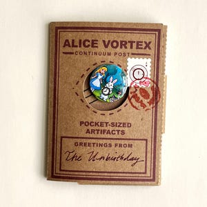 May include: A circular pinback button featuring an illustration of Alice and the White Rabbit with a clock, mounted on a brown cardboard display card labeled ALICE VORTEX CONTINUUM POST POCKET-SIZED ARTIFACTS GREETINGS FROM The Unbirthday.