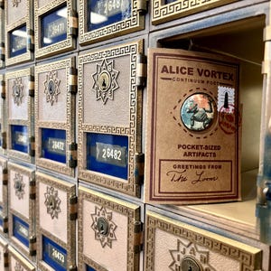 May include: A circular pin featuring a robot illustration attached to a rectangular brown card labelled Alice Vortex Continuum Post Pocket-Sized Artifacts Greetings From The Loom, placed inside a brass-coloured metal post office box unit.