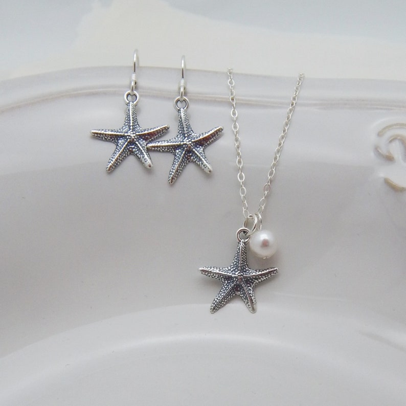 Puede incluir: Un conjunto de collar y pendientes de plata con un colgante de estrella de mar con un acento de perla. El collar tiene una cadena delicada y los pendientes son de estilo colgante.