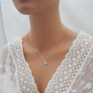 Puede incluir: Un collar de plata con un dije de estrella de mar y un peque&ntilde;o colgante de perla. El collar est&aacute; sobre un fondo de encaje blanco.