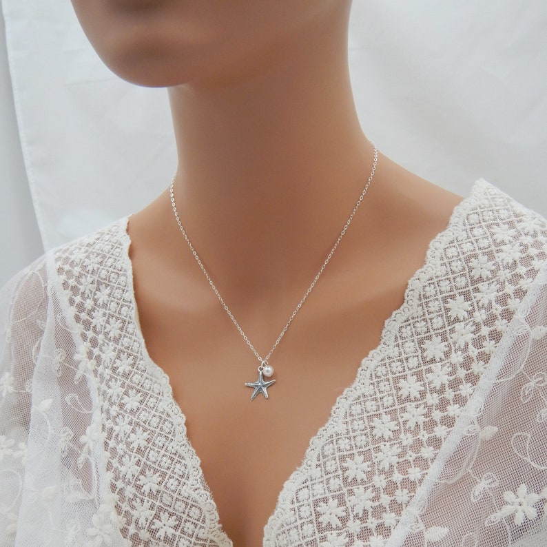 Puede incluir: Un collar de plata con un dije de estrella de mar y un peque&ntilde;o colgante de perla. El collar est&aacute; sobre un fondo de encaje blanco.