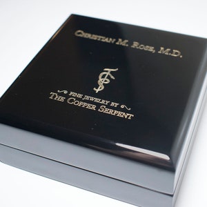 May include: Black jewelry box with gold lettering that reads "Christian M. Rose, M.D. Fine Jewelry by The Copper Serpent".