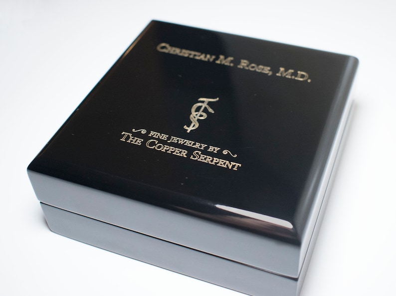 May include: Black jewelry box with gold lettering that reads "Christian M. Rose, M.D. Fine Jewelry by The Copper Serpent".