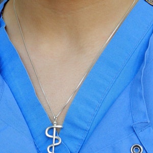 May include: A silver necklace with a caduceus pendant. The necklace is worn by a person wearing a blue shirt.