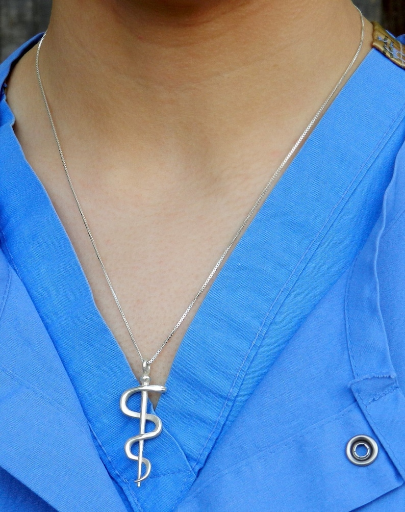 May include: A silver necklace with a caduceus pendant. The necklace is worn by a person wearing a blue shirt.