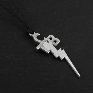 May include: Silver-colored pendant necklace featuring a lightning bolt design with the letters "CB" incorporated into the top. The pendant is suspended from a black cord, creating a striking contrast against the dark background.