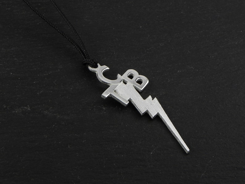 May include: Silver-colored pendant necklace featuring a lightning bolt design with the letters "CB" incorporated into the top. The pendant is suspended from a black cord, creating a striking contrast against the dark background.
