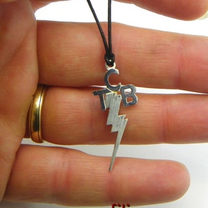 May include: Silver-toned pendant necklace featuring the initials "TCB" above a lightning bolt design. The pendant hangs from a black cord. The necklace is held between fingers.