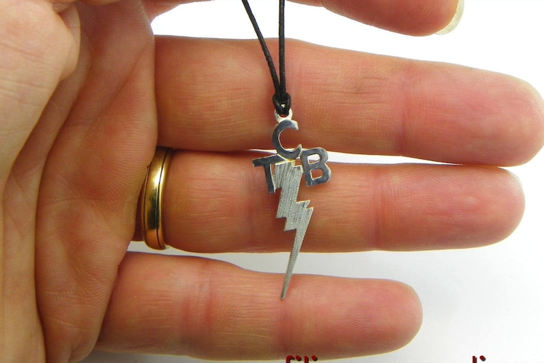 May include: Silver-toned pendant necklace featuring the initials "TCB" above a lightning bolt design. The pendant hangs from a black cord. The necklace is held between fingers.