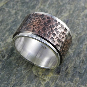 Op de afbeelding: Een draaibare ring van zilver en koper met een gehammerde textuur. De ring bestaat uit twee banden, een van zilver en een van koper, die onafhankelijk van elkaar draaien.