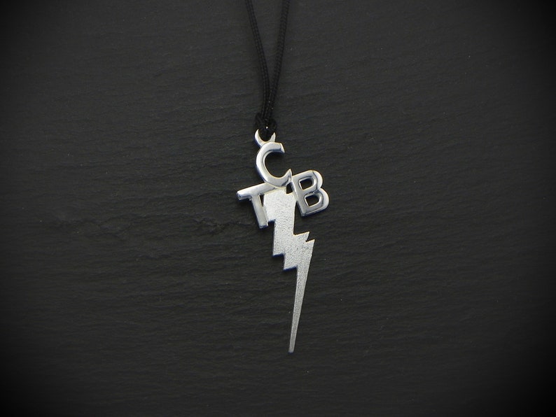 May include: Silver-toned pendant necklace with the letters "TCB" and a lightning bolt design. The pendant hangs from a black cord. The necklace is displayed against a dark, textured background.