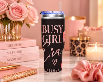 Op de afbeelding: Een zwarte roestvrijstalen beker met een doorzichtig deksel en rietje, voorzien van de roze tekst "BUSY GIRL Era" en een klein hartje, met het woord "BELUVE" in kleine witte letters onderaan gedrukt.
