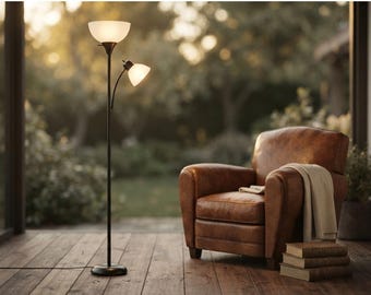 May include: A black metal floor lamp with a large upward-facing frosted glass shade and a smaller adjustable side reading light, positioned next to a brown leather armchair draped with a light-colored throw blanket and a stack of three books.