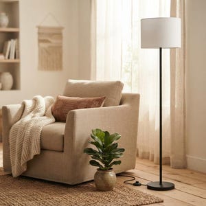 May include: A beige upholstered armchair with a cream-colored knit throw blanket and a rust-colored rectangular pillow. A tall floor lamp with a black metal stand and a white cylindrical shade stands next to a small potted green plant.