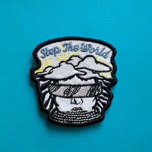 以下が含まれることがあります： 頭の上に雲が描かれた、スタイリッシュな人物の刺繍パッチ。黒と白で刺繍されています。雲の上に「Stop The World」という文字があります。