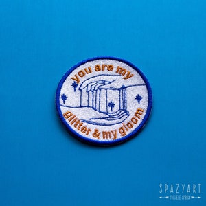 Peut inclure: Un patch brodé bleu et orange avec le texte "You are my glitter & my gloom" et un motif de deux mains tenant une série de lignes verticales.