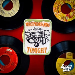 May include: A white embroidered patch with green trim. The patch features a black and white illustration of a face with the text "WHAT I'M DREAMING TONIGHT" in orange and black.