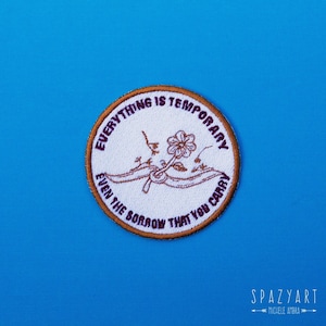Op de afbeelding: Een witte geborduurde patch met een bruine rand. De patch heeft een bloem met een stengel en bladeren, en de tekst "Alles is tijdelijk, zelfs het verdriet dat je draagt".