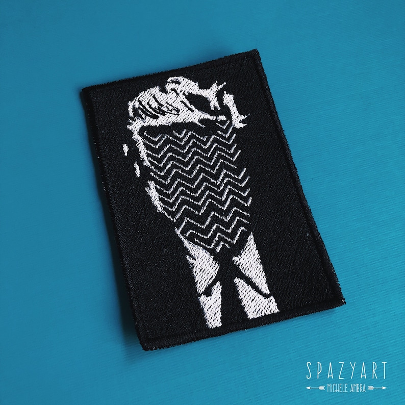 Peut inclure: Patch brod&eacute; noir et blanc repr&eacute;sentant la silhouette d'une personne avec un motif en zigzag sur la poitrine. Le patch est sur un fond bleu.