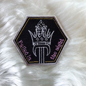 May include: A black hexagonal patch with a white embroidered crown and the text "Felled in the night" in pink.