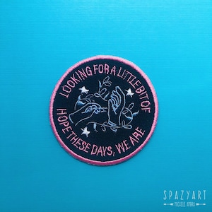 May include: A black embroidered patch with pink stitching. The patch features two hands reaching towards a small plant with a star on each side. The text around the image reads "Looking for a little bit of hope these days, we are".