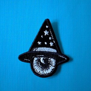 May include: Black and white embroidered patch of an eye wearing a black wizard hat with white stars.
