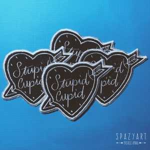 Könnte beinhalten: Fünf schwarze Filzpatches in Herzform mit weißen Nähten und einem Pfeil-Design. Jeder Patch sagt "Stupid Cupid" in weißer Kursivschrift.