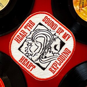 May include: A white embroidered patch with red stitching. The patch features a black and white illustration of a heart with a wave coming out of it. The text "Hear the Sound of My Heart Exploding" is written around the illustration.