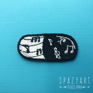Puede incluir: Parche bordado en blanco y negro con forma de píldora con notas musicales sobre un fondo azul. El texto "Spazyart Michele Ambra" está debajo del parche.
