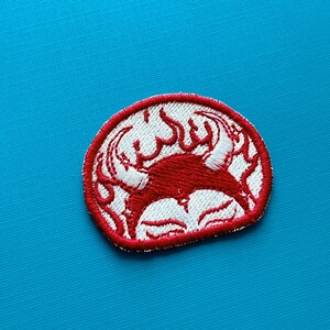 Pu&ograve; includere: Toppa ricamata con un design di granchio rosso e dettagli bianchi, incorniciata da un bordo rosso. Il granchio &egrave; stilizzato con fiamme e una forma circolare. La toppa &egrave; su uno sfondo blu.