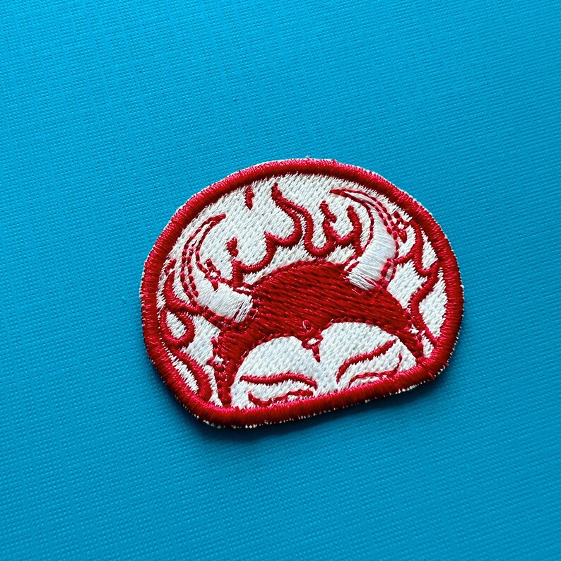 Pu&ograve; includere: Toppa ricamata con un design di granchio rosso e dettagli bianchi, incorniciata da un bordo rosso. Il granchio &egrave; stilizzato con fiamme e una forma circolare. La toppa &egrave; su uno sfondo blu.