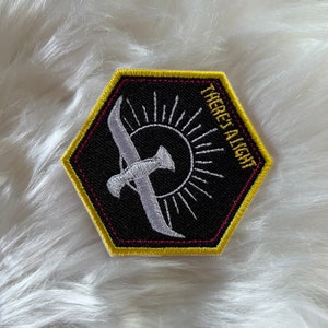 May include: A black hexagonal patch with a white embroidered design of a bird flying towards a sun with rays. The text "THERE'S A LIGHT" is embroidered in white on the right side of the patch. The patch has a yellow border and a red inner border.