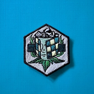 Puede incluir: Parche bordado hexagonal. El diseño presenta una cara con un patrón de tablero de ajedrez, hojas verdes y un borde blanco y negro. El parche está sobre un fondo azul claro.