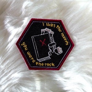 May include: A black hexagonal embroidered patch with a red X and the text "I was the match you were the rock" in gold thread.
