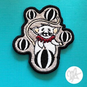 May include: Embroidered patch featuring a white mouse juggling black and white striped balls. The mouse wears a red ruffled collar and is set against a gray background with a black border. The patch is on a turquoise background.