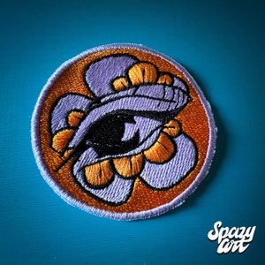 May include: A round embroidered patch with an orange background and a purple flower with an eye in the centre. The eye is black with a white pupil. The flower has orange petals.