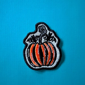Puede incluir: Un parche de calabaza bordado en blanco y negro con una calabaza naranja con rayas blancas y negras.